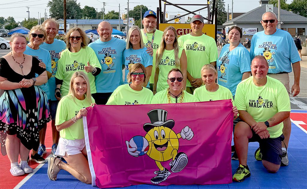 Group photo of smiling people wearing matching bright shirts, holding a colorful banner outdoors.
