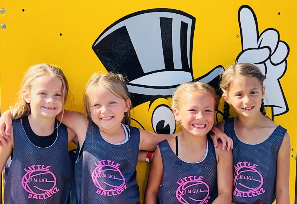 Four smiling young girls in matching sports tank tops stand in front of a colorful cartoon backdrop.